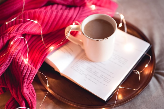 Tasse Kaffee bleibt auf offenem Buch auf rundem Holztablett mit Strickpullover und Weihnachtsbeleuchtung Guten Morgen