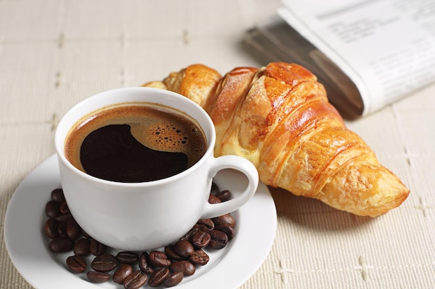 Tasse Kaffee, Croissant und Zeitung auf verdeckter Tischdecke