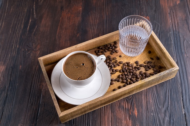 Tasse Kaffee, Kaffeebohnen und Glas Wasser in einer Holzkiste auf einem dunklen Holz