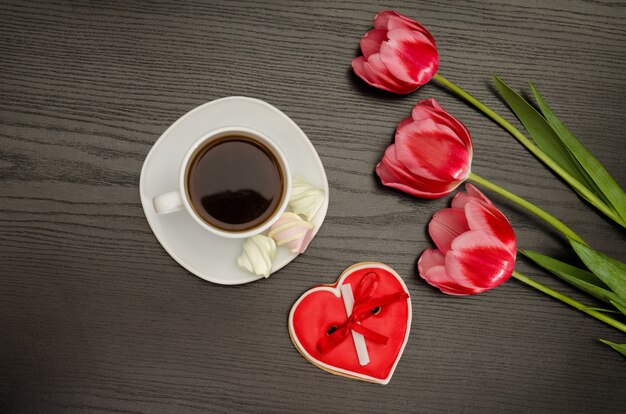 Tasse Kaffee, Marshmallows und Lebkuchen in Herzform, drei rosa Tulpen. Schwarzer Hintergrund.