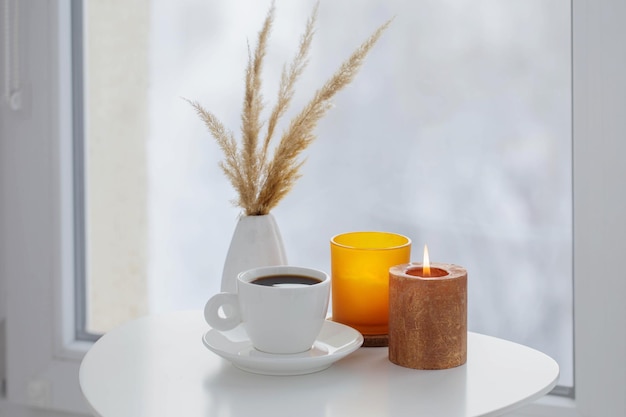 Tasse Kaffee mit brennenden Kerzen auf weißem Tisch am Fenster im Winter