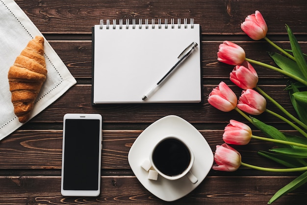 Tasse Kaffee mit einem Hörnchen zum Frühstück auf dem Tisch verziert mit einem Blumenstrauß von rosa Tulpen und von Smartphone
