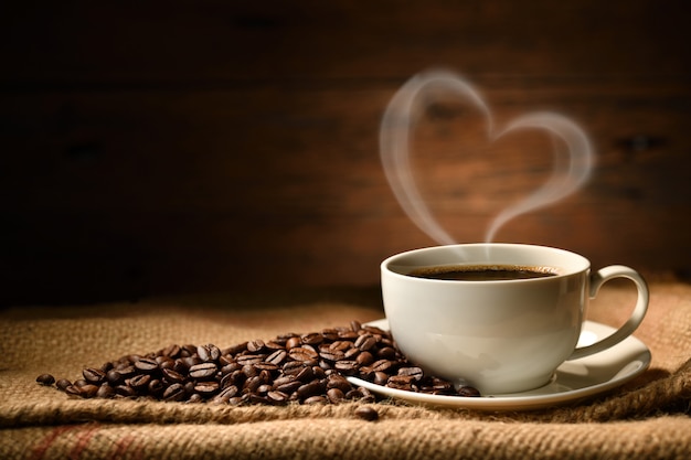 Tasse Kaffee mit Herzformrauch und Kaffeebohnen auf Leinensack auf altem hölzernem Hintergrund