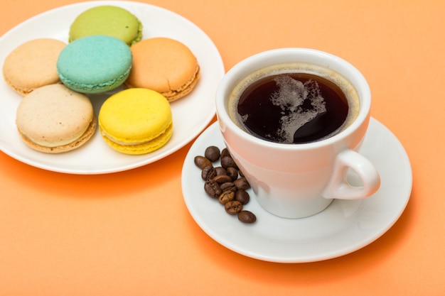 Tasse Kaffee mit Kaffeebohnen auf Untertasse und köstlichen Macarons-Kuchen unterschiedlicher Farbe auf weißem Porzellanteller mit pfirsichfarbenem Hintergrund. Selektiver Fokus auf Tasse