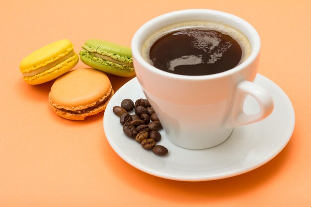 Tasse Kaffee mit Kaffeebohnen auf weißer Untertasse und leckeren Macarons-Kuchen in verschiedenen Farben mit pfirsichfarbenem Hintergrund