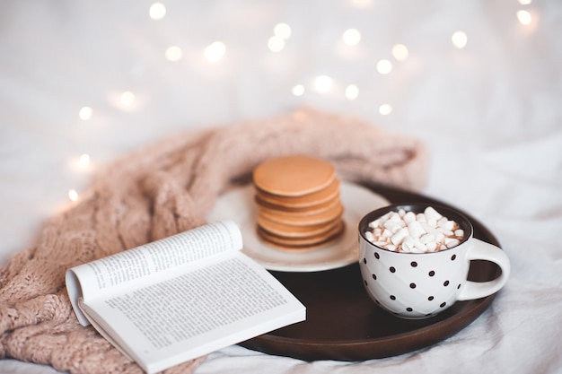 Tasse Kaffee mit Marshmallow Aufenthalt auf Holztablett mit offenem Buch und Pfannkuchen im Bett über Weihnachtsbeleuchtung Guten Morgen Frühstück