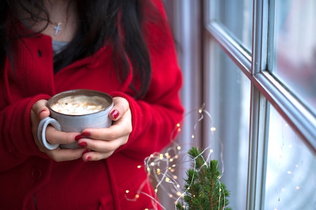 Tasse Kaffee mit Marshmallow in den Händen einer Frau in einem roten Pullover, in der Nähe des Fensters