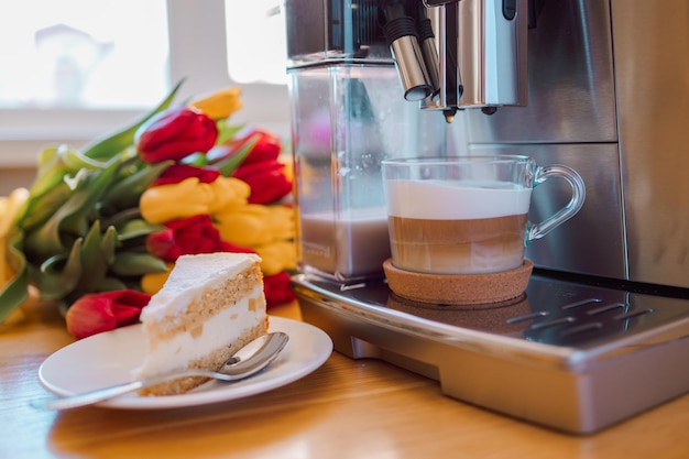 Tasse Kaffee mit Milch Stück Kuchen und Tulpen Blumen auf Küchentisch aus Holz Frisch gebrühter Cappuccino Espresso in der Kaffeemaschine zum Frühstück Stilvolle Inneneinrichtung