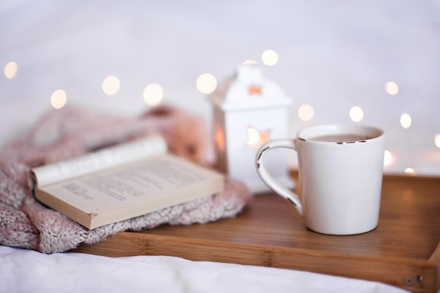 Tasse Kaffee mit offenem Buch und gestricktem Pullover auf Holztablett hautnah über Weihnachtsbeleuchtung Guten Morgen Ferienzeit