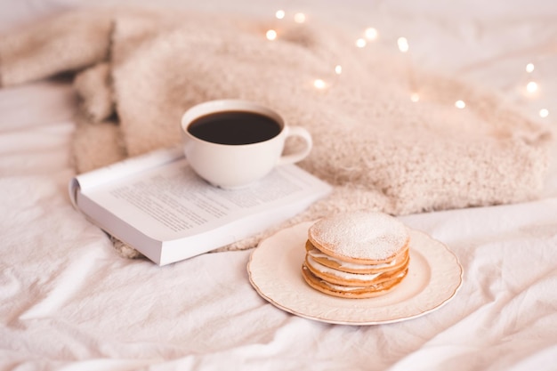 Tasse Kaffee mit Pfannkuchen und offenem Buch über Strickpullover im Bett. Warme und gemütliche Atmosphäre. Wintersaison. Guten Morgen.