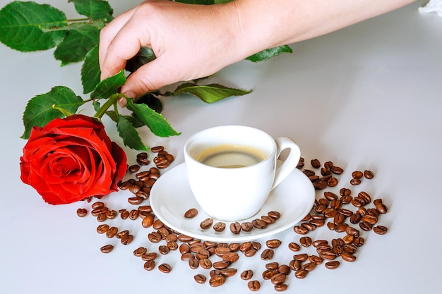 Tasse Kaffee mit Rose auf weißem Hintergrund. Kaffeekörner sind auf dem Tisch verstreut.