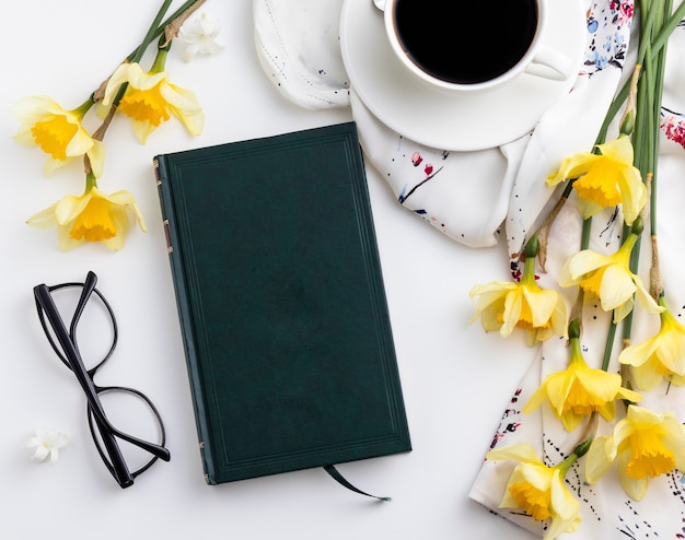 Tasse Kaffee mit schönen gelben Blumen, einem schwarzen Notizbuch und einer Brille auf weißem Hintergrund