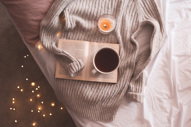 Tasse Kaffee oder schwarzer Tee auf offenem Buch mit brennender Kerze und gestricktem grauen Pullover im Bett über leuchtenden Weihnachtslichtern. Winterferienzeit. Ansicht von oben.