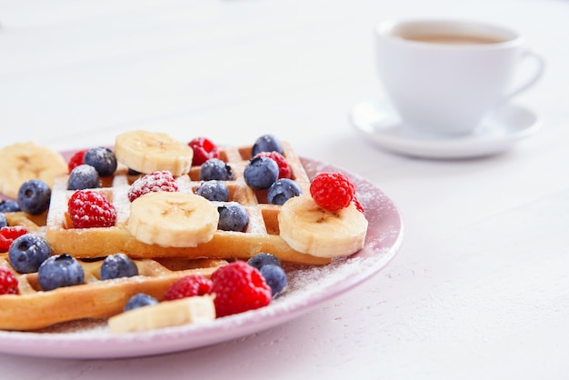 Tasse Kaffee und belgische Waffeln mit Blaubeeren, Himbeeren, Bananen und Zuckerpulver auf weißem Hintergrund. Konzept von leckerem und gesundem Essen.