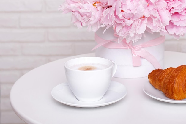 Tasse Kaffee und Croissant mit Pfingstrosenblumen auf weißem Tisch