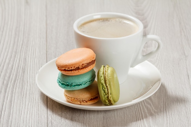 Tasse Kaffee und köstliche Macarons-Kuchen in verschiedenen Farben auf weißem Porzellanteller mit grauem Holzhintergrund