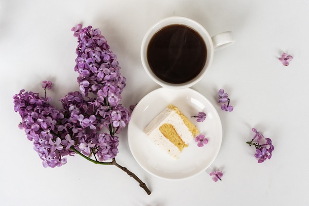 Tasse Kaffee und Kuchenhörner ausStillleben mit einem Strauß Flieder auf einem weißen Tisch, eine Tasse Kaffee, ein Teller mit einem Stück Kuchen. Internationaler Frauentag, 8. März