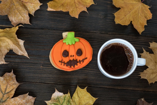 Tasse Kaffee und Lebkuchen als Kürbislaterne auf Holztisch neben Herbstlaub Halloween