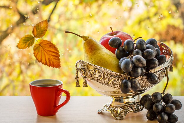 Tasse Kaffee und Metallvase mit gelber Birne, Weintraube und Apfel. Trockenes Blatt auf Fensterglas mit Wassertropfen und Herbstbäumen im Hintergrund.