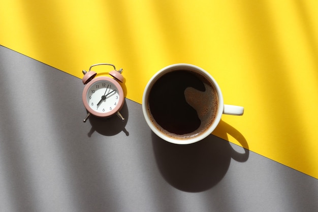 Tasse Kaffee und Wecker auf trendigem gelbem und grauem Hintergrund mit tiefen Schatten, die Morgenkonzept aufwachen