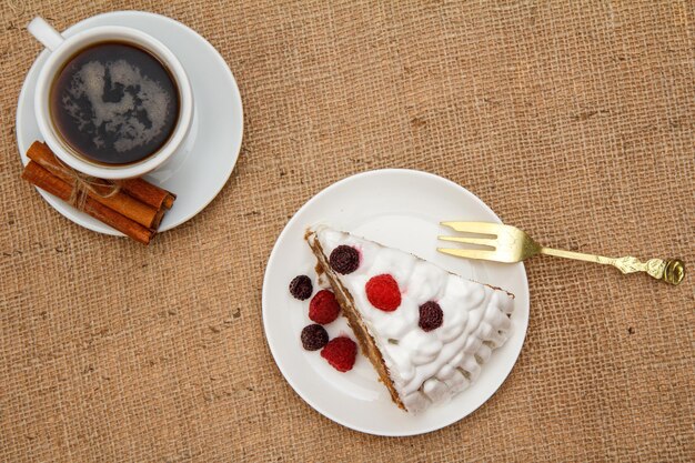 Tasse Kaffee, Zimt, Gabel und Kekskuchen mit Schlagsahne und Himbeeren auf dem Tisch mit Sackleinen. Ansicht von oben.