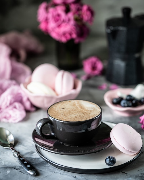 Tasse Kaffee zum Frühstück mit Marshmallows in Form von Makronenkuchen in einer schwarzen Schüssel auf einem dunklen Tisch und mit Blumen in einem Glas