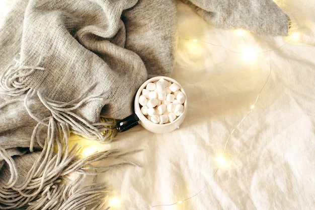 Tasse Kakao mit Marshmallows, Schal, Scheinlichter auf weißem Bettlaken. Weihnachten, Winter, Neujahrskomposition. Flache Lage, Ansicht von oben, Kopienraum.