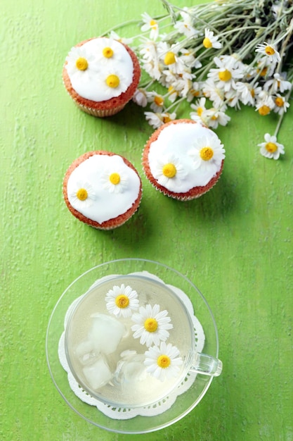 Tasse Kamillentee mit Kamillenblüten und leckeren Muffins auf farbigem Holzhintergrund