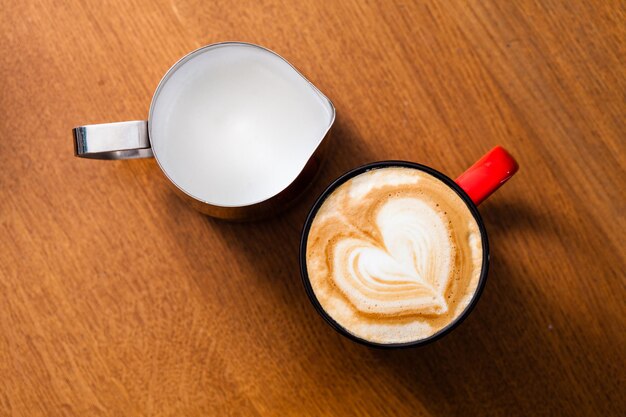 Tasse Latte Art Cappuccino und Krug mit Milchschaum auf Holztisch in Draufsicht