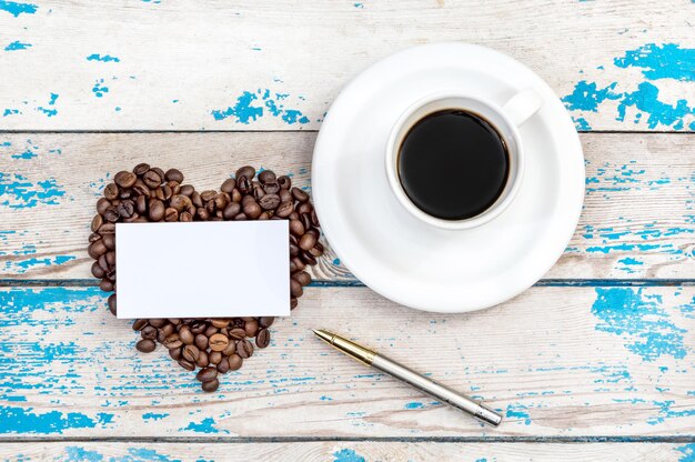 Tasse mit Kaffee mit Herz aus Kaffeebohnen Visitenkarte und Stift auf Draufsicht aus Holzhintergrund