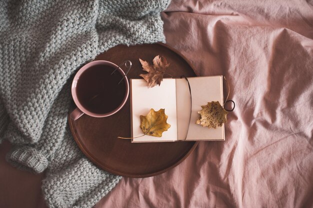 Tasse schwarzen Tee mit offenem Notizbuch auf Holztablett und Strickpullover im Bett aus nächster Nähe. Herbstsaison. Guten Morgen. Frühstückszeit.