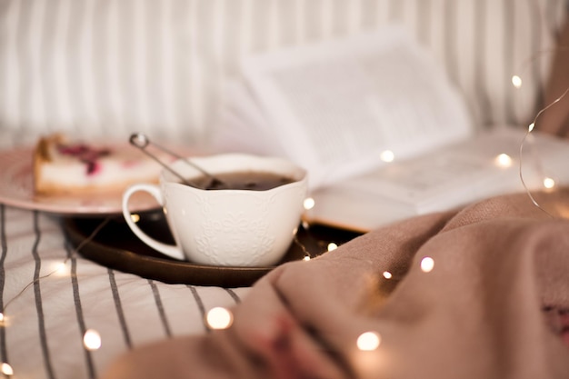 Tasse schwarzen Tee über offenem Buch und Lichtern im Hintergrund in der Bettnahaufnahme. Guten Morgen. Frühstück.