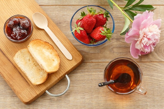 Tasse Tee, hausgemachte Erdbeermarmelade und frische Beeren in Glasschalen, Holzlöffel, Toast auf Schneidebrett und Pfingstrose. Ansicht von oben.