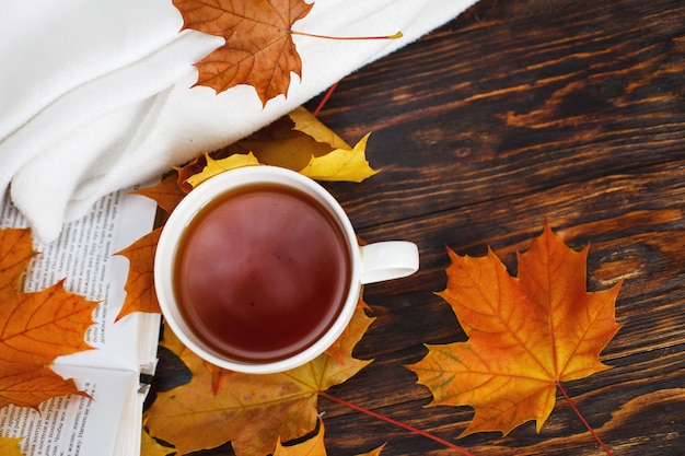 Tasse Tee mit goldenem Herbstlaub kariert und Buch auf einem hölzernen Hintergrund