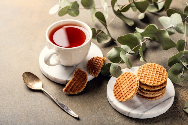 Tasse Tee mit Mini-Stroopwafel, Sirupwaffelkeksen und Eukalyptuszweigen auf heller Oberfläche