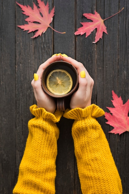 Tasse Tee mit Zitrone in einer weiblichen Hand. Hölzerner Hintergrund, Herbstlaub. Ansicht von oben