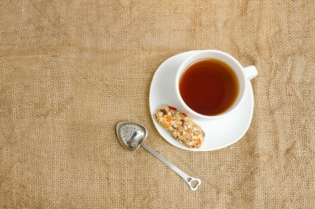 Tasse Tee, Müsliriegel und Teesieb auf Sackleinen. Ansicht von oben