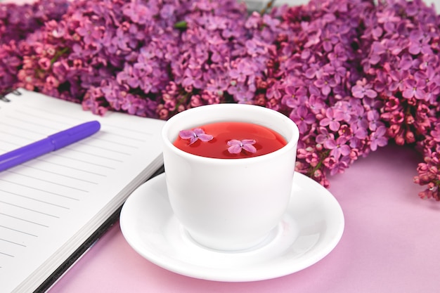 Tasse Tee und Niederlassungen des Frühlinges blüht blühende Flieder des Blumenstraußes auf purpurroter Tabelle.