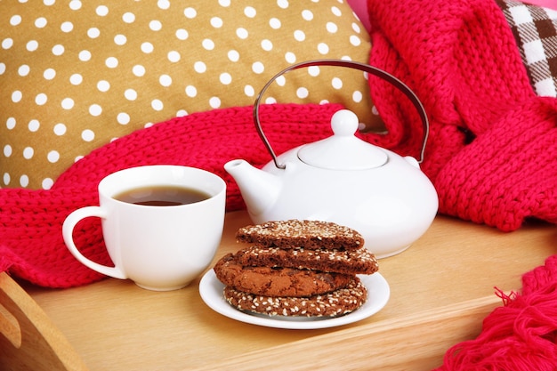 Tasse und Teekanne mit Keksen auf Tablett und Schal auf dem Bett aus nächster Nähe