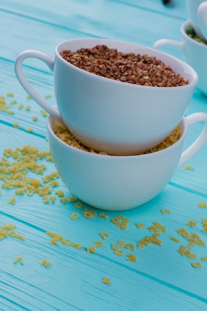 Tasse voller Buchweizengrütze, die über einer anderen Tasse steht. Blauer Holztischhintergrund.