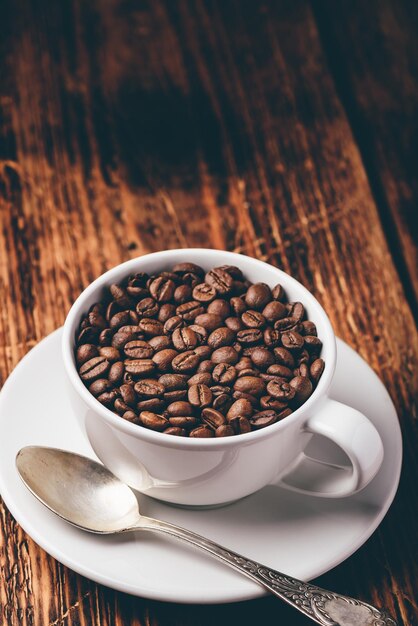 Tasse voller gerösteter Kaffeebohnen auf Untertasse über Holztisch