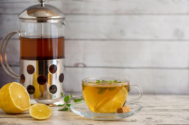 Tasse Zitronentee mit Rohrzucker, französischer Presse und Zitronenscheiben auf hellem Holztisch