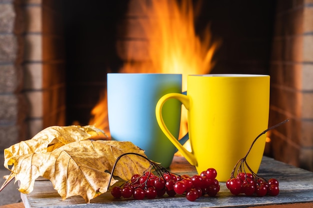 Tassen Tee oder Kaffee und gelbes Herbstlaub in der Nähe eines gemütlichen Kamins in einem Landhaus