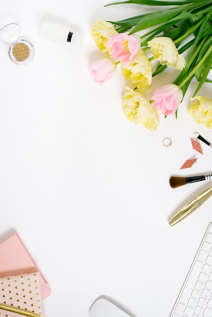 Tastatur auf weißem Hintergrund mit Tulpenblumen, Notizblöcken, Kosmetik, Schmuck und Flat-Lay-Zubehör. Ansicht von oben. Das Konzept eines weiblichen Freiberuflerbüros.