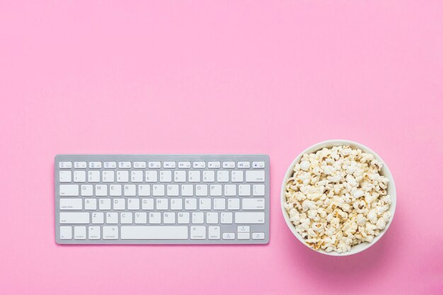 Tastatur und Schüssel Popcorn auf einem Rosa. Konzept zum Ansehen von Filmen, TV-Shows und Shows online. Flache Lage, Draufsicht.