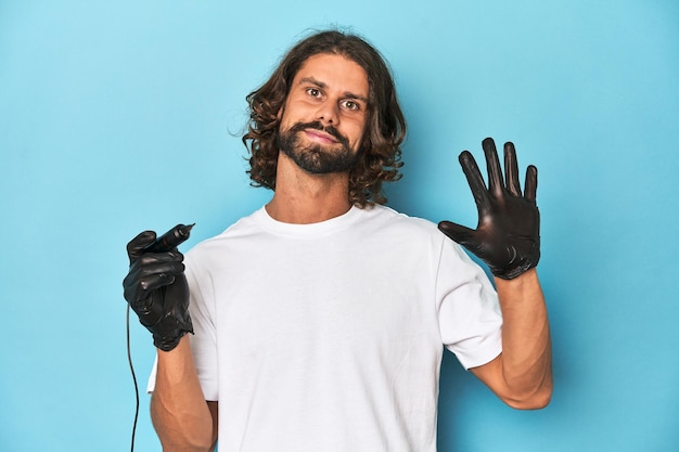 Tatuador barbudo con máquina de tatuaje en el trabajo sonriendo alegremente mostrando el número cinco con los dedos