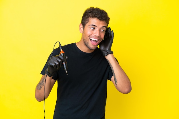 Tatuador brasileño aislado de fondo amarillo escuchando algo poniendo la mano en la oreja