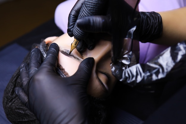 Un tatuador con guantes negros realiza maquillaje permanente de cejas. una mano sostiene la cabeza del cliente, la otra trabaja con una máquina de tatuar