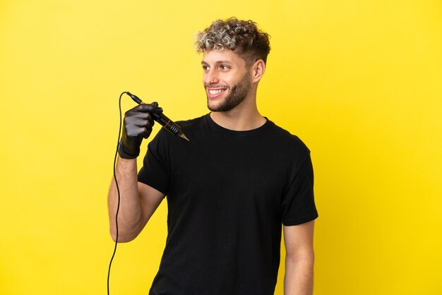 Tatuador homem caucasiano isolado em um fundo amarelo, olhando para o lado e sorrindo