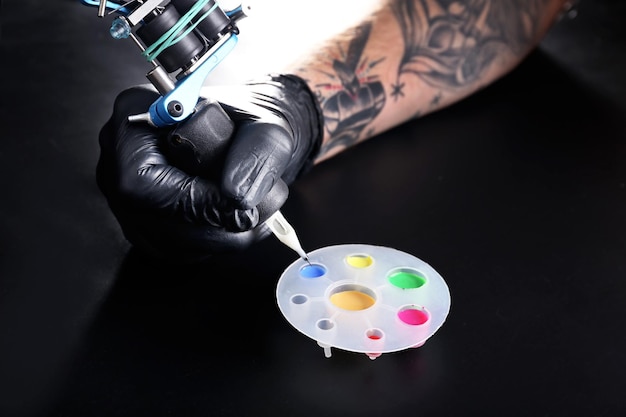 Tatuador mano en guante médico con máquina de tatuaje y tintas de colores sobre fondo negro
